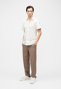 Vīrietis tērpies baltā īsroku krekliņā ar maziem daudzkrāsainiem punktiem, brūnās brīvā stilā biksēs un baltās sporta kurpēs, stāvot ar rokām kabatās.