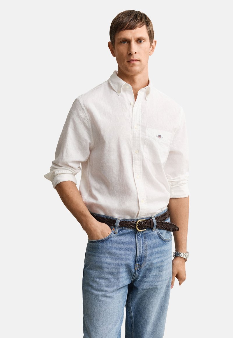 Jeune homme portant une chemise blanche à boutons avec manches retroussées, un jean bleu, une ceinture brune tressée et une montre-bracelet argentée, debout avec une main dans la poche.