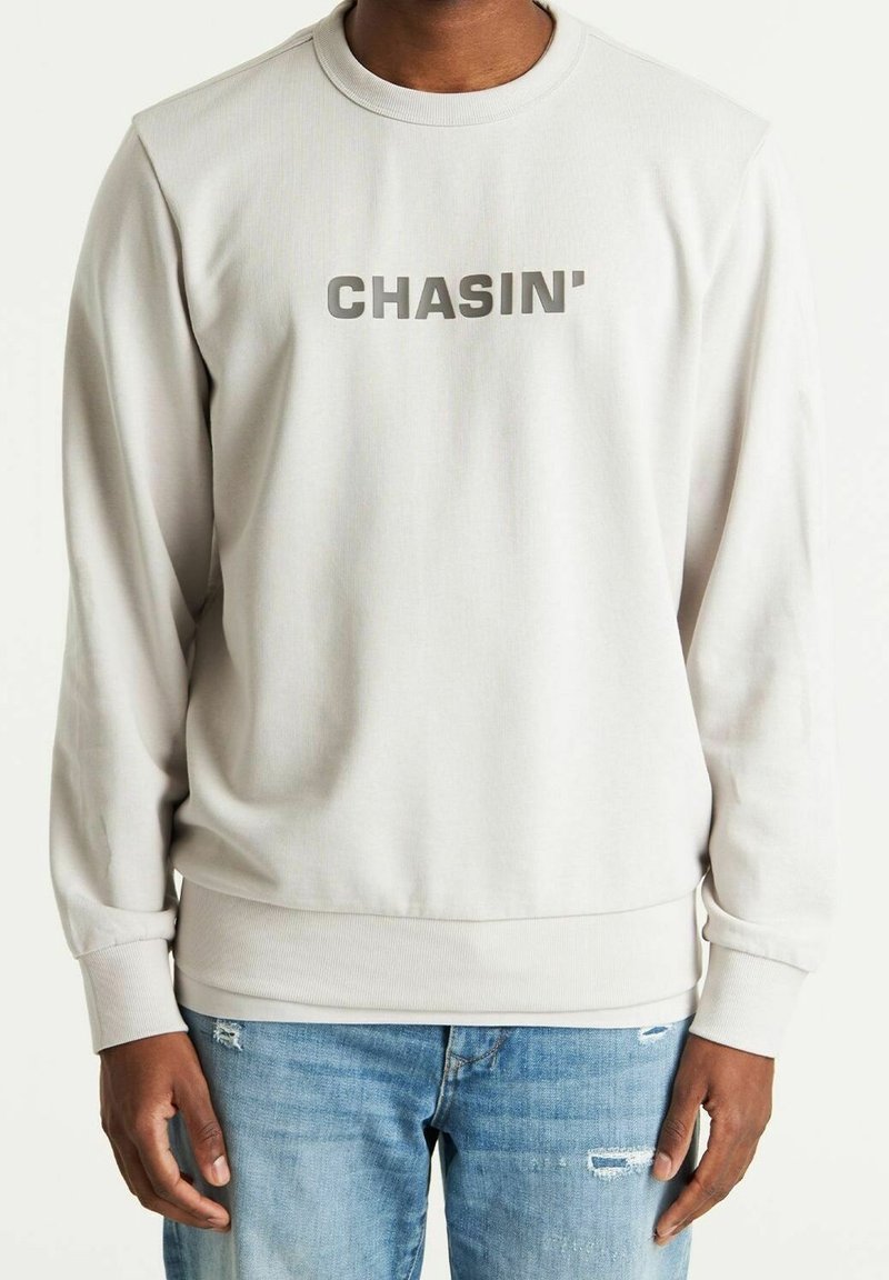Personne portant un sweat-shirt beige clair avec le texte "CHASIN'" sur la poitrine et un jean bleu déchiré, debout sur fond blanc.