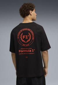 Mustat puuvillapaita rennolla leikkauksella. Takana punainen graafinen kuva, jossa on laakerinlehvädesign ja "F1"-logo, juhlistaen Formula 1:n 75-vuotispäivää.