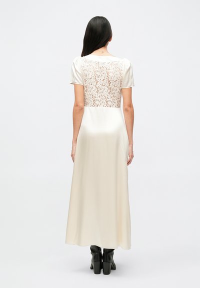 Ilga, kreminės spalvos suknelė trumpomis rankovėmis; nugaros dalyje nėriniuota panelė; priekyje lygi satino medžiaga; sluoksniuotas apatinis kraštas; derinama su juodais aukštakulniais batais.