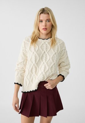 Cream knitted sweater with a cable knit pattern, contrasting black trim, paired with a burgundy pleated skirt. Textured, loose-fit design.