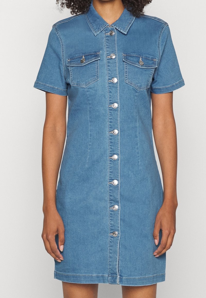 Kvinde iført en kortærmet blå denimkjole med sølvfarvede knapper foran og to brystlommer med klap.