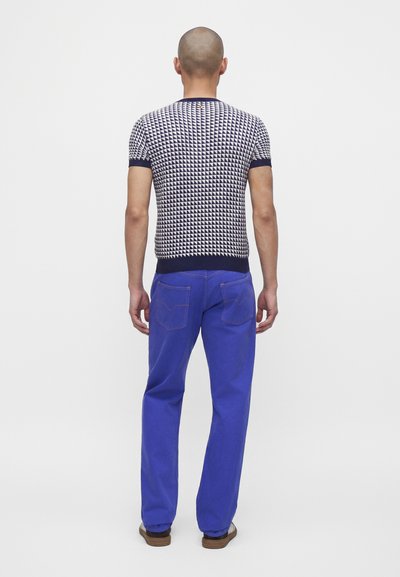 Homme à la tête rasée portant une chemise noire et blanche à manches courtes à motifs et un pantalon bleu vif, debout de dos sur un fond uni.