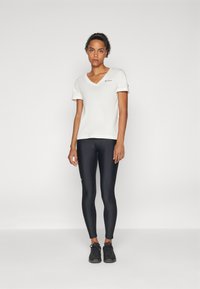 T-shirt bianco a V con maniche corte e un logo sottile, abbinato a leggings neri aderenti e scarpe da ginnastica nere. Texture lisce.