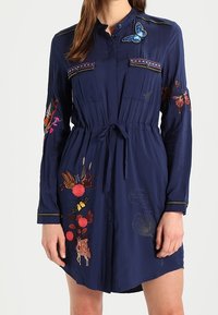 Mujer con un vestido azul marino de mangas largas, con mariposas y flores bordadas de colores, y un lazo en la cintura.