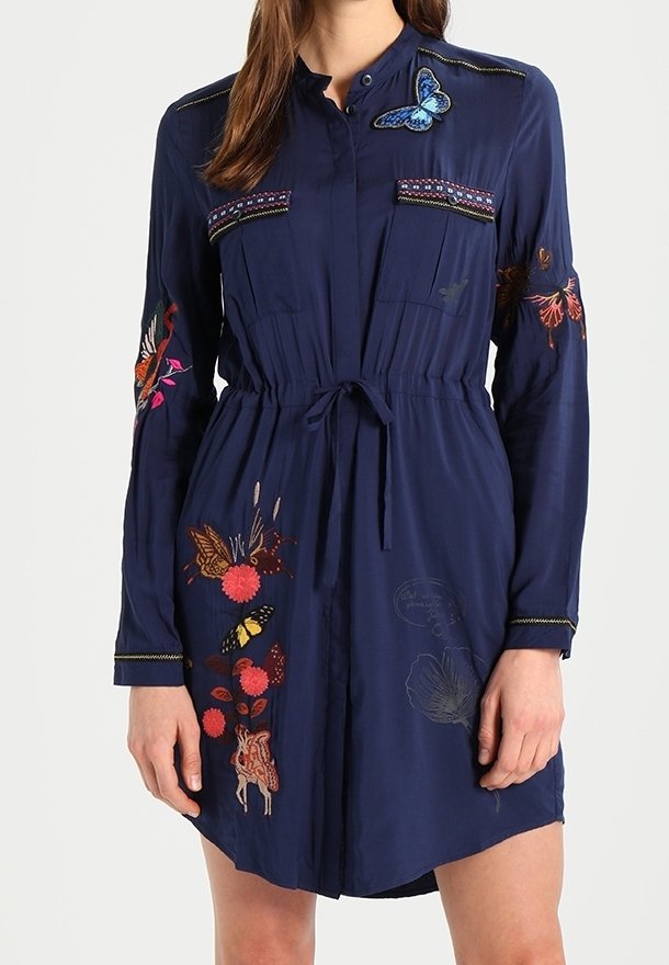 Mujer con un vestido azul marino de mangas largas, con mariposas y flores bordadas de colores, y un lazo en la cintura.