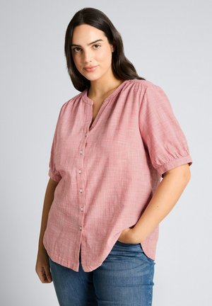 Frau mit dunklen Haaren, die eine rosa kurzärmelige Bluse mit Knöpfen und blaue Jeans trägt, steht mit einer Hand in der Tasche vor einem grauen Hintergrund.