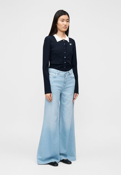 Σκούτερ μπλε ναυτικό με ριμπ υφή, λευκό γιακα και κουμπιά, συνδυασμένο με ανοιχτό μπλε τζιν παντελόνι με φαρδύ πόδι. Μαύρα παπούτσια ολοκληρώνουν την εμφάνιση σε ουδέτερο φόντο.