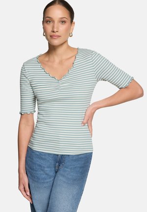 Femme portant un haut à manches courtes rayé bleu clair et blanc avec des bords festonnés et un jean bleu, posant avec une main sur la hanche.