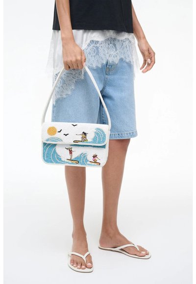 Femme tenant un sac à main blanc avec des surfeurs, des vagues, le soleil et des oiseaux brodés, portant un haut bordé de dentelle, un short en jean et des sandales blanches.
