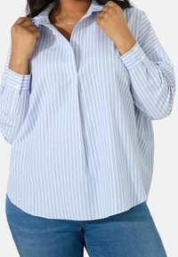 Donna che indossa una camicia a righe verticali blu chiaro e bianca con jeans blu, mentre tiene il colletto della camicia con entrambe le mani.