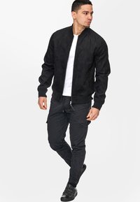 Schwarze Kunstwildleder-Bomberjacke mit Reißverschluss, gerippten Manschetten und Kragen. Kombiniert mit schwarzen Cargo-Hosen und schwarzen Schuhen.