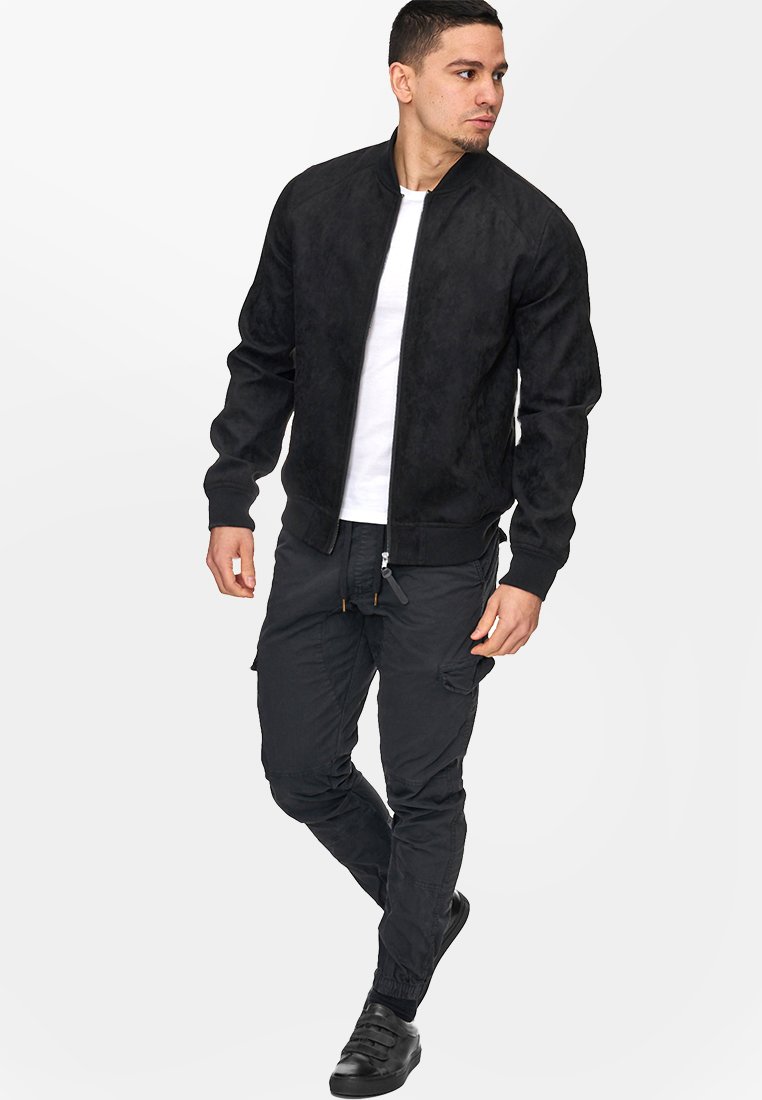 Veste bomber en faux suede noir avec fermeture éclair à l'avant, poignets et col côtelés. Associée à un pantalon cargo noir et des chaussures noires.
