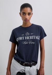 Camiseta de algodón azul marino con mangas cortas, con texto blanco: "SPORT HERITAGE" y "Scalpers Company." Diseño estándar de cuello redondo.