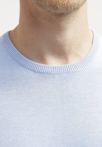 Collo e parte bassa del viso di una persona con una leggera barba, che indossa un maglione lavorato a maglia di colore azzurro chiaro con un collo a coste.