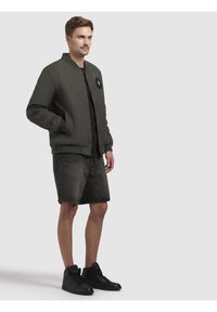 Olive rohelise bomber-jakk ümmarguse patchiga, lühikeste varrukatega, ribakäistega, milles on mustad denimsärgud ja mustad kõrge säärega spordijalatsid.