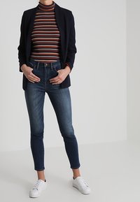 Blazer navy, cuello alto a rayas en rojo, azul y naranja, jeans slim-fit de color azul oscuro y zapatillas blancas. Look moderno y ajustado.