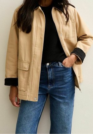 Femme portant une veste beige par-dessus un haut noir et un jean bleu, avec une main dans la poche et de longs cheveux foncés.