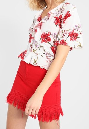 Vrouw draagt een witte blouse met rood bloemenprint en een rode minirok met franjes aan de zoom, poserend tegen een effen achtergrond.