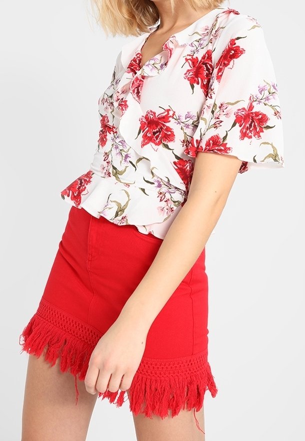 Femme portant un chemisier blanc à imprimé floral rouge et une mini-jupe rouge à ourlet à franges, posant devant un fond uni.