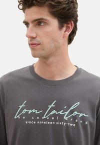 Dark gray long-sleeve t-shirt featuring a printed logo in light blue and white. Round neckline and soft, smooth fabric texture.