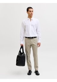 Άνδρας που φοράει λευκό πουκάμισο με κουμπιά και μπεζ παντελόνια, κρατώντας μαύρη τσάντα duffel με το δεξί χέρι, στέκεται σε απλό ανοιχτόχρωμο φόντο.