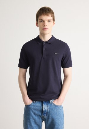 Polo bleu marine en coton, avec un col classique, une patte de boutonnage à trois boutons et un petit patch logo noir sur la poitrine.