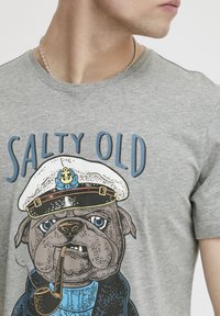 Grå t-skjorte med et grafisk motiv av en bulldog iført en hvit kapteinslue, som holder en pipe, med teksten "SALTY OLD" i blått.