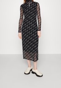 Robe noire en maille à manches longues avec un motif répétitif de texte blanc, col montant, et longueur mi-mollet. Associée à des sabots crème à semelle épaisse.