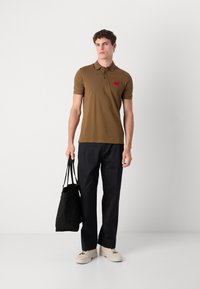 Cămașă polo maro cu mâneci scurte, având o patch cu logo roșu. Asortată cu pantaloni negri și o geantă tote neagră. Pantofii sunt bej.