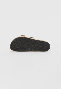 Des chaussures avec une semelle texturée noire et un dessus beige, présentant deux sangles réglables. Le design est plat et simple.