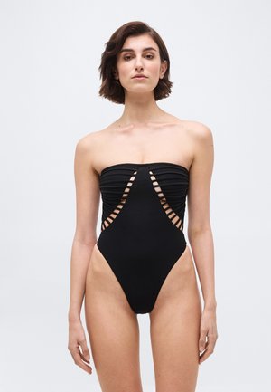 Donna che indossa un costume da bagno nero senza spalline con un design a scaletta a taglio sul davanti e sui lati, in piedi contro uno sfondo chiaro e uniforme.