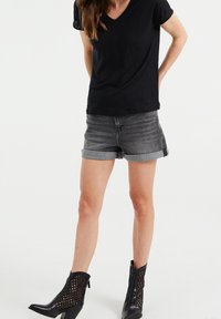 Kvinna klädd i svart kortärmad topp, rullade grå denimshorts och svarta mesh stövletter, stående mot en vit bakgrund.