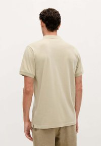 Camiseta polo beige de manga corta, hecha de una tela suave y texturizada. Presenta un corte recto y un cuello clásico. Se muestra vista trasera.