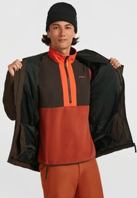 Fleece pullover met bruine en feloranje panelen, half-zip ontwerp, gedragen onder een donkere buitenjas. Bevat een gebreide zwarte beanie.