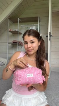 Trousse de maquillage matelassée rose avec motif floral et logo, dotée d'une fermeture éclair et d'une forme compacte et arrondie. Tissu texturé avec une finition brillante.