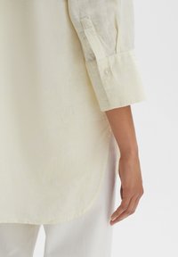 Light yellow striped shirt with a relaxed fit, featuring a button cuff and a curved hem, complemented by a smooth, textured fabric.