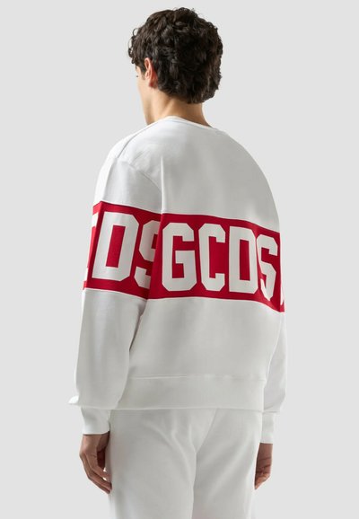 Person som bär vit sweatshirt och byxor, sett bakifrån, med ett brett rött band och stora vita bokstäver "GCDS" över övre delen av ryggen och ärmarna.