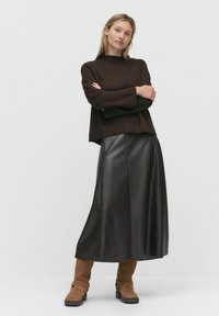Pull en tricot marron, jupe en cuir noire et bottes en cuir brun jusqu'aux genoux avec boucles. Le mannequin se tient les bras croisés devant un fond neutre.