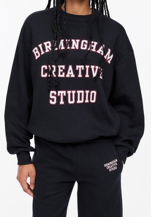 Personne portant un sweat-shirt et un pantalon noirs avec un texte blanc et rose indiquant "Birmingham Creative Studio" sur les deux vêtements.