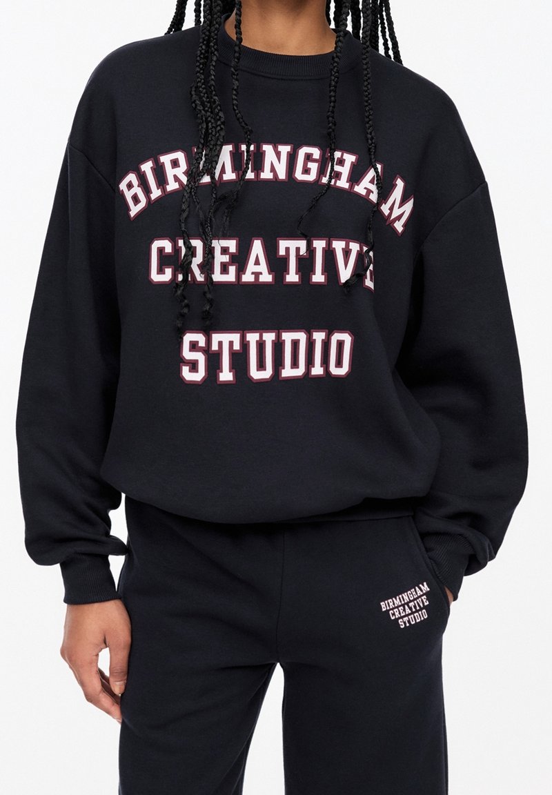 Personne portant un sweat-shirt et un pantalon noirs avec un texte blanc et rose indiquant "Birmingham Creative Studio" sur les deux vêtements.