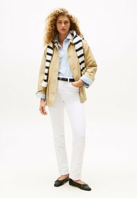 Beige regenjas, lichtblauw gestreept shirt, witte broek en zwarte platte schoenen. Gestreepte sjaal over de schouders gedrapeerd. Neutrale achtergrond.