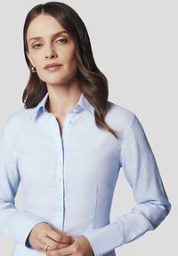 Jasnoniebieska koszula z długim rękawem wykonana z lnu, z kołnierzykiem i zapięciem na guziki z przodu, wyeksponowana na neutralnym tle.