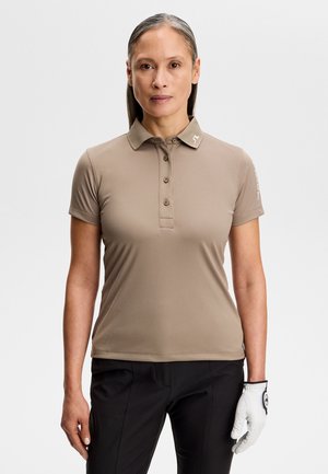 Kvinde iført en beige kortærmet poloshirt, sorte bukser og en hvid golfhandske på venstre hånd, stående mod en ensfarvet baggrund.