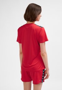 Maglietta sportiva rossa a maniche corte, con accenti neri sulle spalle e una texture liscia. Indossata con pantaloncini rossi abbinati.