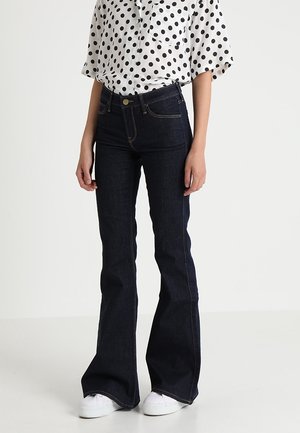 Mujer con vaqueros oscuros acampanados, zapatillas blancas y una camiseta blanca de manga corta con lunares negros, de pie frente a un fondo liso.