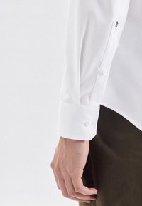 Weißes Hemd mit Knopfleiste und strukturiertem Finish, ausgestattet mit einem sauberen Bund mit zwei Knöpfen und einem kleinen gestickten Detail am Ärmel.