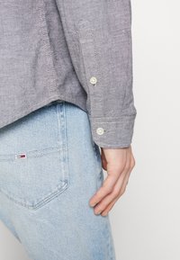 Gray button-up shirt with a textured fabric, featuring single-button cuffs, paired with light blue denim jeans, showing rear pocket detail.