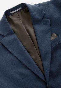 Tengerészkék zakó behajtott gallérral, látható barna mintás béléssel és hozzáillő barna díszzsebkendővel a mellzsebben.